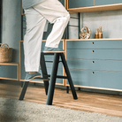 Een persoon in een witte broek en slippers staat op een Hasegawa Lucano Ladder 2 Step Zwart van Hasegawa in een moderne, op Japan geïnspireerde woonkamer met blauwe kasten, houten planken en decoratieve voorwerpen op de achtergrond.