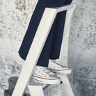 Een persoon in een donkere broek en witte sneakers met rode en blauwe accenten staat op een Hasegawa Lucano Ladder 3 Step Wit van Hasegawa, ontworpen in Japan, tegen een lichtgekleurde muur met textuur.