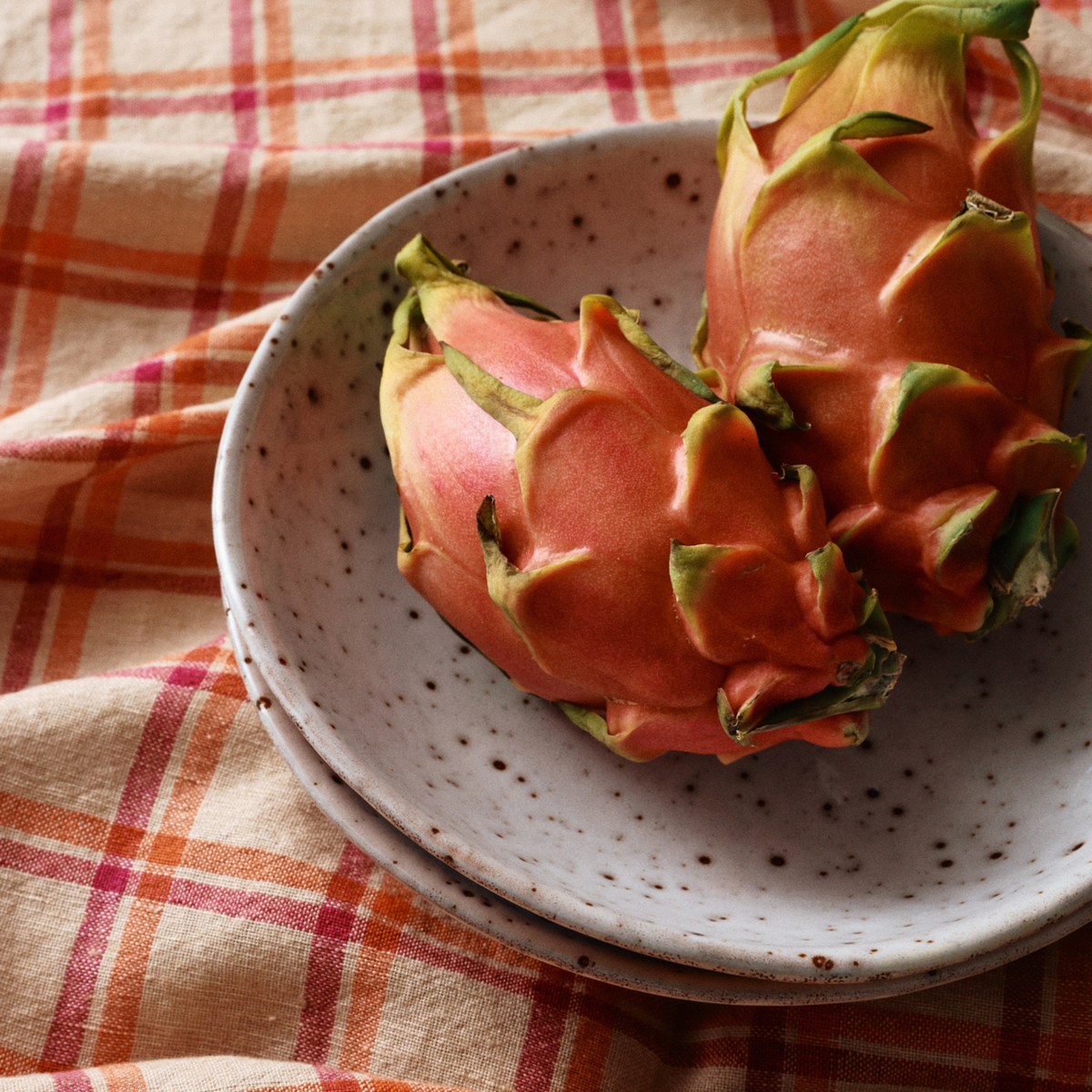 Twee hele drakenvruchten met rozerode schil en groen gestipte schubben rusten op een HK Living 70s Ceramics Curry Kom Hibiscus bord, op een geruite oranje en beige stoffen achtergrond.