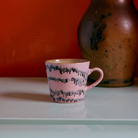 Een roze HK Living 70s Ceramics Cappuccinokop Bubble met een zwart golvend patroon rust op een lichtgroen oppervlak, met een grote bruine vaas op de achtergrond tegen een oranje muur.