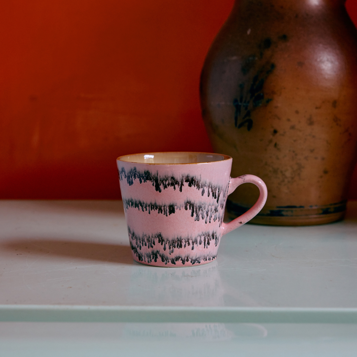 Een roze HK Living 70s Ceramics Cappuccinokop Bubble met een zwart golvend patroon rust op een lichtgroen oppervlak, met een grote bruine vaas op de achtergrond tegen een oranje muur.