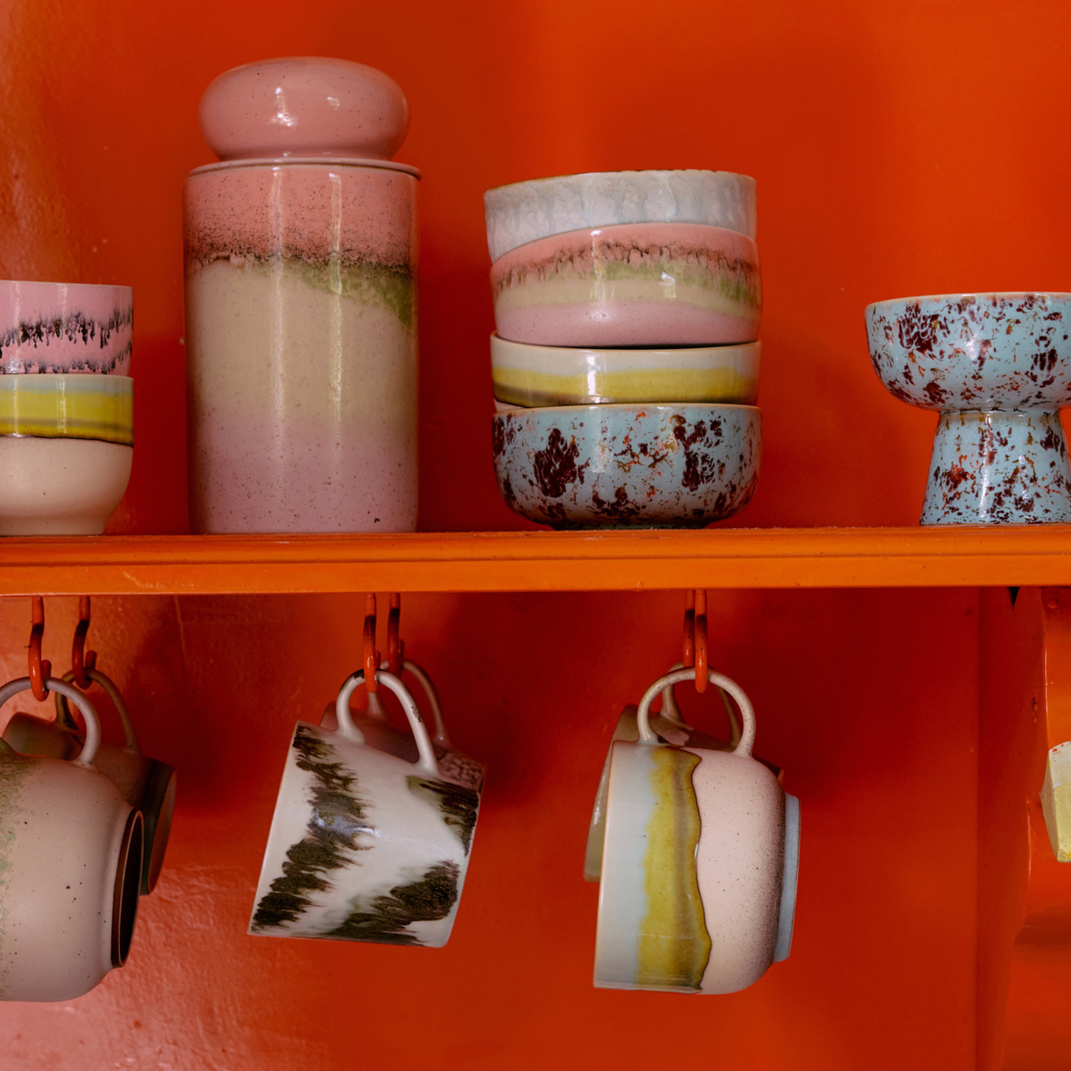 HK Living 70s Ceramics Cappuccinokop Tide mokken met artistieke patronen hangen aan hun handvatten, terwijl bijpassende kommen en een bus met reactief glazuur erboven staan, allemaal tegen een levendige oranje plank.