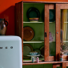 Een vintage keuken met een lichtblauwe SMEG koelkast, een houten kast met verschillende borden, kommen en HK Living 70s Ceramics Pasta Kom Patina 2 st. tegen groene en oranje muren.