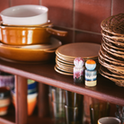 Gestapelde bruine schalen, kommen en rieten borden staan op een houten plank. Aan de voorkant staan twee HK Living 70s Ceramics Peper- en Zoutvaatje Stargaze shakers, met retro tafelaccessoires en meer borden op de achtergrond.