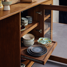 Een houten keukenkastje met glazen deuren gaat open en laat HK Living 70s Ceramics Pasta Kom Petal 2 st. kommen zien, netjes gerangschikt op planken, naast borden en mokken in aardetinten van HK Living.