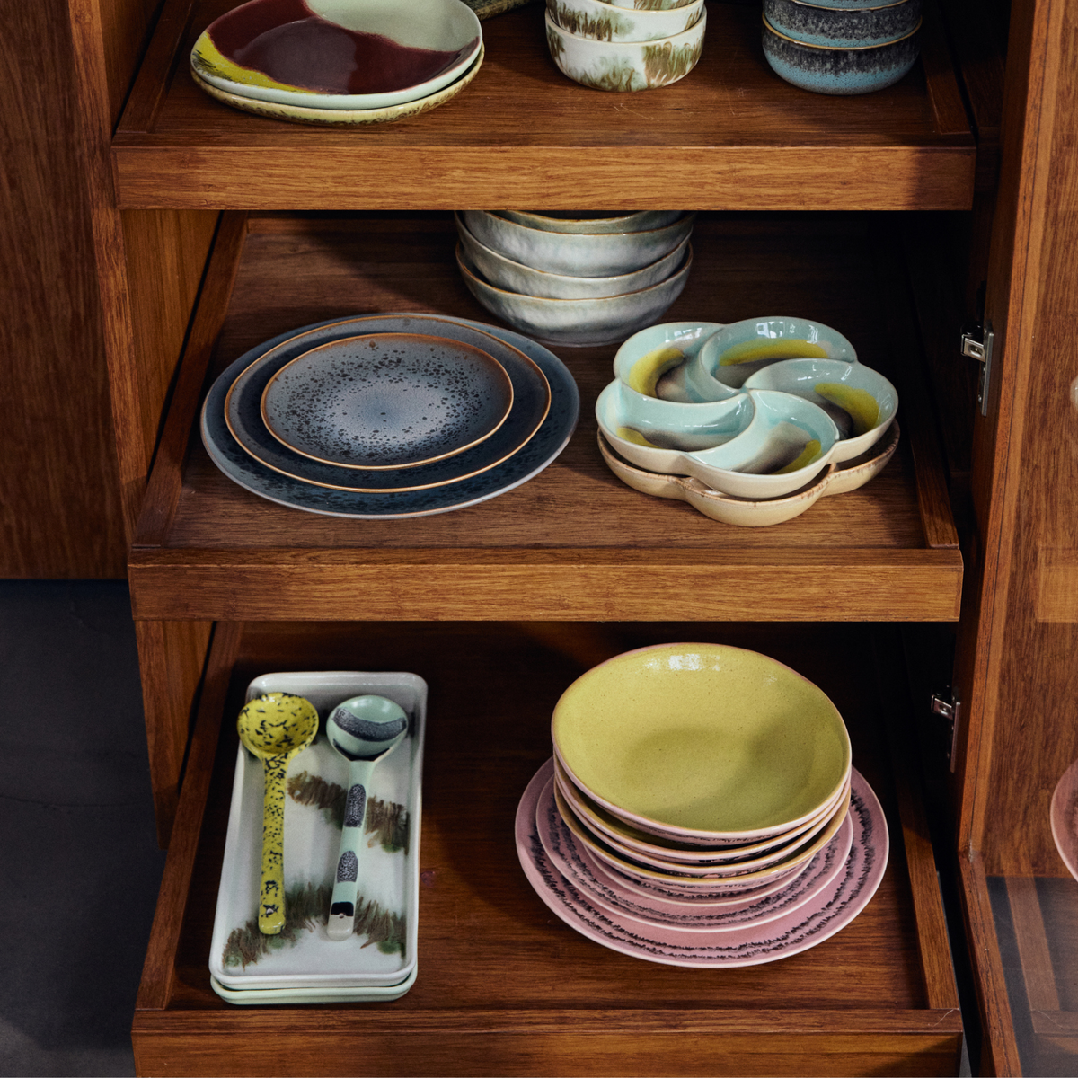 Een houten kast met open planken toont nette sets HK Living 70s Ceramics Pasta Kom Petal 2 st. met hun kleurrijke patronen en design.