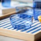 Een set van twee Lyngby Glas Torino Kom Blauw D12 kommen rust op een houten dienblad met een blauw-wit gestreepte doek, terwijl vage Lyngby Glas Torino glazen en een citroenpartje op de achtergrond verschijnen.