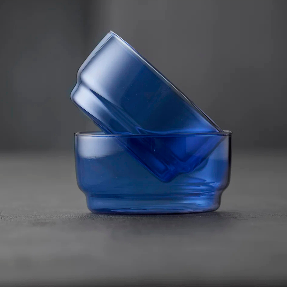 Twee doorschijnende kommen (set van 2) van Lyngby Glas Torino Kom Blauw D12 staan in een hoek in elkaar op een glad grijs oppervlak en tonen het elegante ontwerp dat kenmerkend is voor Lyngby Glas.