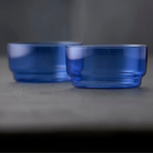 Twee Lyngby Glas Torino Kom Blauw D12 kommen van Lyngby Glas rusten op een grijs oppervlak; één kom van borosilicaatglas is scherp in beeld aan de voorkant, terwijl de andere een beetje onscherp is op de achtergrond.
