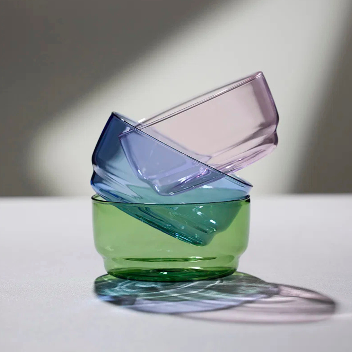 Twee doorschijnend groene Lyngby Glas Torino kommen (D12) zijn diagonaal gestapeld op een wit oppervlak in zachte verlichting, waarbij hun borosilicaatglas kleurrijke schaduwen werpt. Merk: Lyngby Glas. Product: Lyngby Glas Torino Kom Groen D12 2 st.