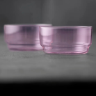 Twee transparante, lichtroze Lyngby Glas Torino Kom Roze D12 kommen (set van 2) rusten op een grijs oppervlak, de ene in focus terwijl de andere zachtjes naar de achtergrond verdwijnt. Merk: Lyngby Glas.