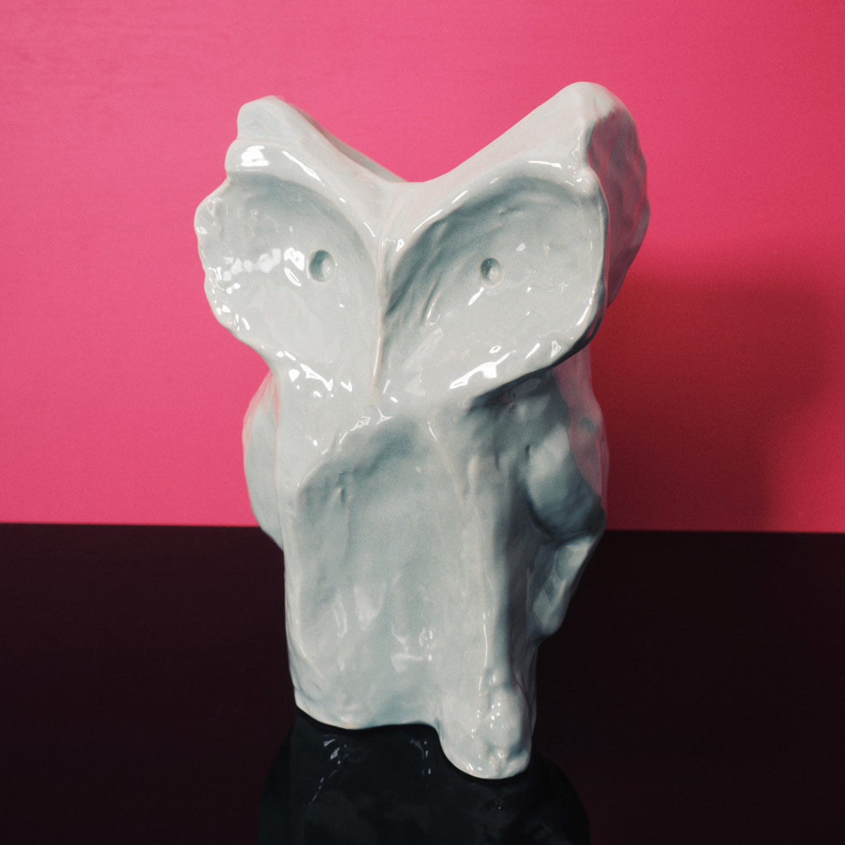 De Serax Marni After Midnight Uil Vaas Lichtblauw S, een abstracte vaas van keramiek in de vorm van een uil, staat op een zwart oppervlak tegen een levendige roze achtergrond.