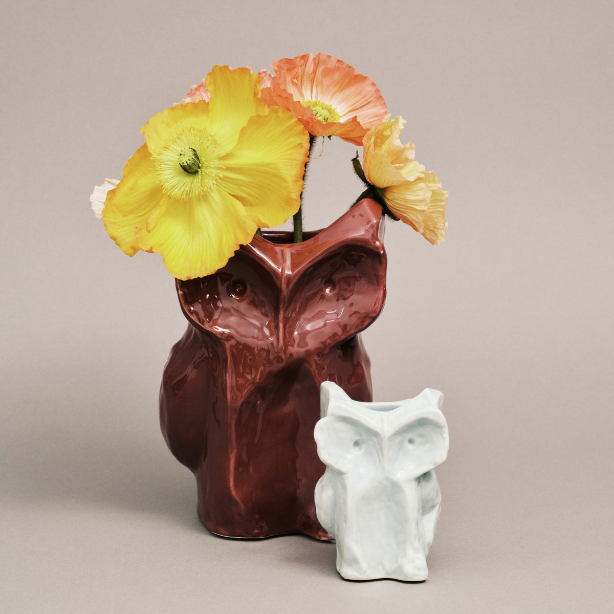 Een glanzende lichtblauwe Serax Marni After Midnight Uil Vaas Lichtblauw XS, in de vorm van een uil, houdt oranje en gele klaproosbloemen vast. Aan de voorkant zit een kleiner wit uilenbeeldje, beide tegen een effen beige achtergrond.