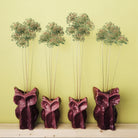 Vier Serax Marni After Midnight Uil Vaas Aubergine S, elk gevuld met lange groene alliumstengels, zijn gerangschikt op een lichte houten plank tegen een lichtgele achtergrond - een verwijzing naar de speelse esthetiek van Marni design.