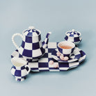 Het Serax Wouters & Hendrix Fête Des Bêtes Melkkannetje Blauw Wit wordt getoond als onderdeel van een blauw-wit geblokt Serax theeservies, gerangschikt met roze snoepjes op het schoteltje tegen een lichtblauwe achtergrond.