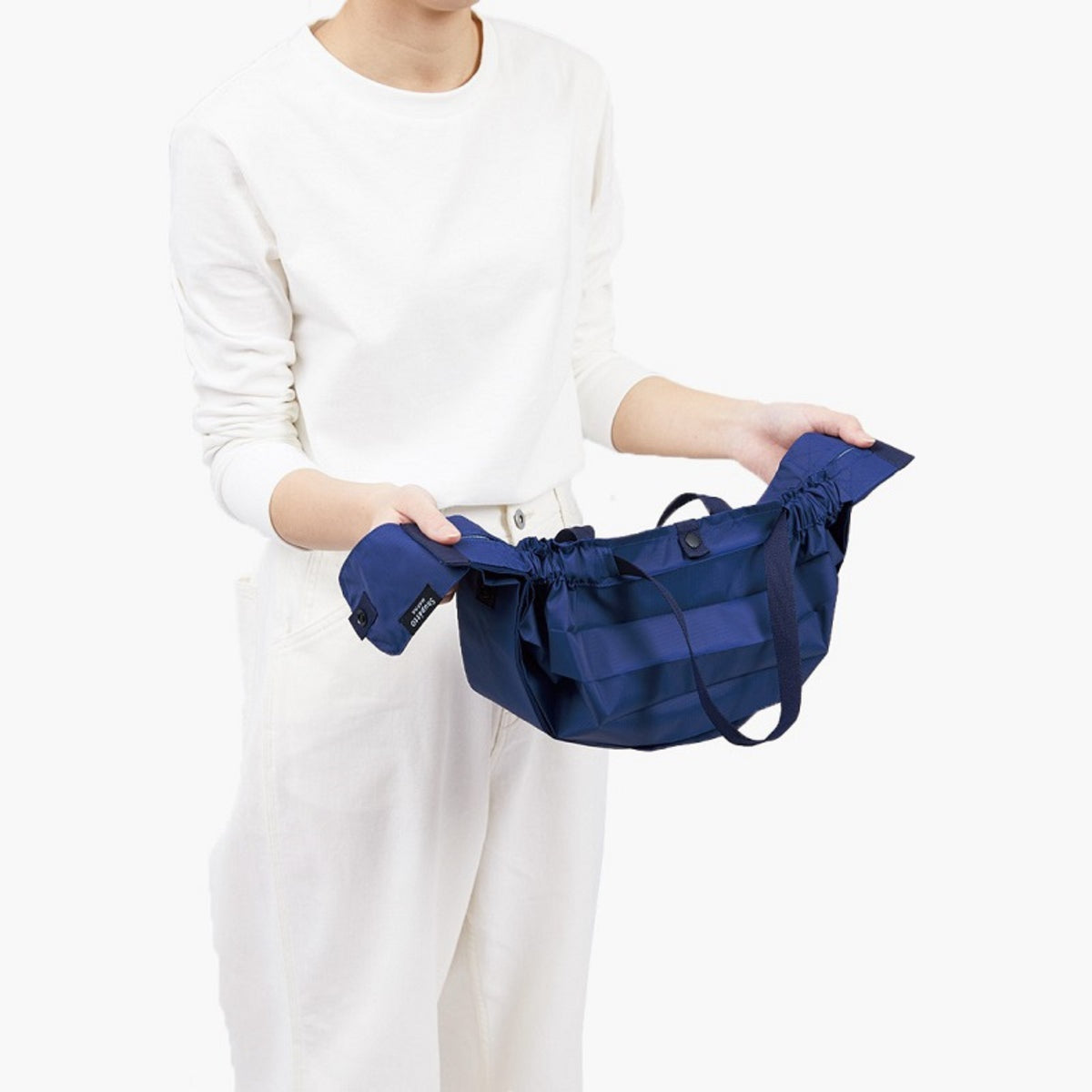 Een persoon in een wit shirt met lange mouwen en een witte broek houdt een open Shupatto Opvouwbare Boodschappentas Forest Groen M tegen een effen witte achtergrond.
