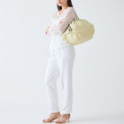 Een vrouw staat tegen een effen witte achtergrond en draagt een transparante witte blouse en broek. Ze draagt de Shupatto Opvouwbare Boodschappentas Cashewnoten Beige M over haar schouder met haar armen gekruist.
