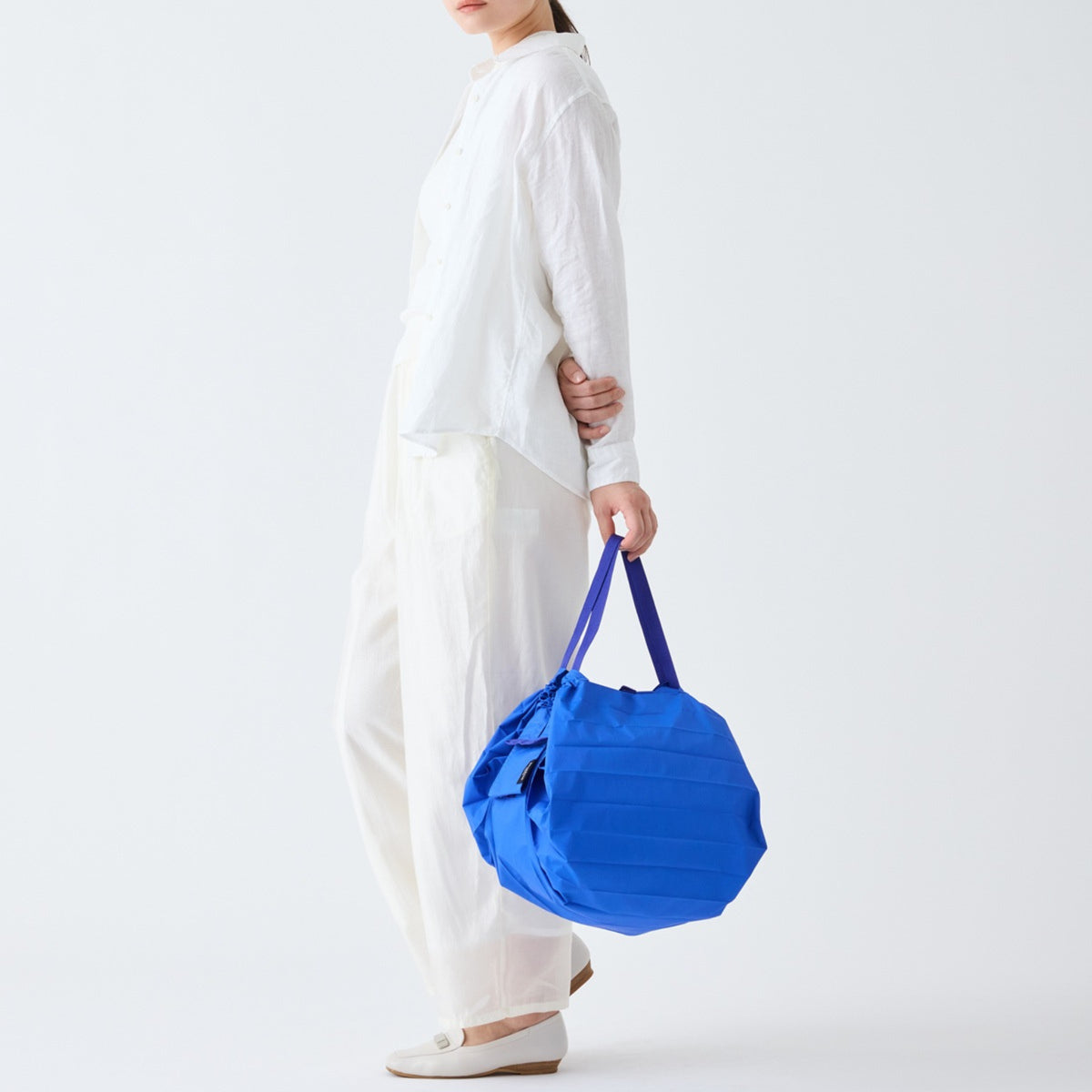 Een persoon in losse witte kleding houdt een Shupatto Opvouwbare Boodschappentas Clear Sky Blauw M van Shupatto in de linkerhand tegen een effen witte achtergrond, een beetje opzij kijkend met alleen het ondergezicht zichtbaar.