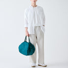 Een persoon in een wit t-shirt en lichtbeige broek houdt de Shupatto Opvouwbare Boodschappentas Donkergroen M vast en maakt zijn minimalistische look af met witte sneakers. Deze praktische, lichtgewicht tas van Shupatto is perfect voor dagelijks gebruik.