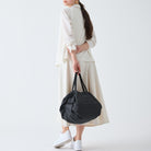 Een vrouw in een off-white jurk met lange mouwen en witte sneakers staat met haar rug naar haar toe gekeerd en houdt een Shupatto Opvouwbare Boodschappentas Jet Zwart M van Shupatto vast tegen een effen witte achtergrond.