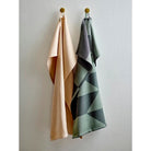 Twee handdoeken hangen naast elkaar aan ronde haken. De linker is effen beige, terwijl de rechter een Södahl Twist Theedoek Groen is met een grijs en groen geometrisch patroon, wat Scandinavische keukenlinnen flair brengt aan de witte muur.