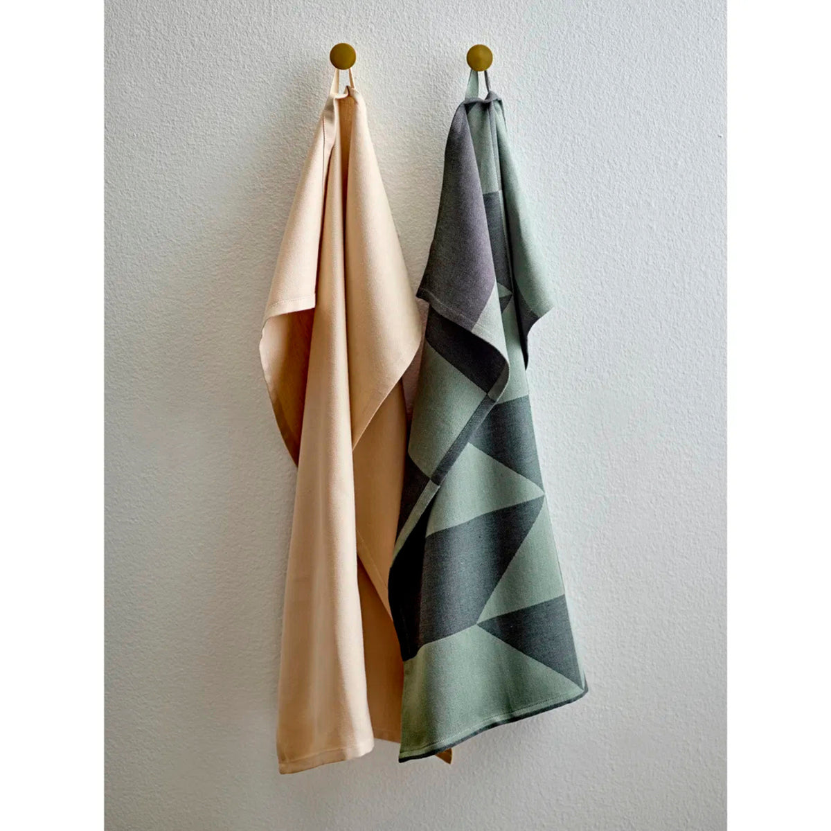 Twee handdoeken hangen naast elkaar aan ronde haken. De linker is effen beige, terwijl de rechter een Södahl Twist Theedoek Groen is met een grijs en groen geometrisch patroon, wat Scandinavische keukenlinnen flair brengt aan de witte muur.